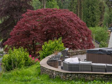An outdoor seating area with gray wicker furniture and a hot tub near trees and bushes at Hazelseat House Graythwaite near Ulverston