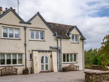 Exterior of a cream-colored house with multiple windows wooden benches and stone wall at Hazelseat House Graythwaite near Ulverston