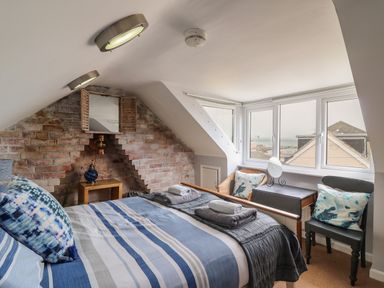 A bedroom with a bed with striped bedding a wooden side table exposed brick wall a wicker chair a small desk with a mirror and two chairs near a window at Harbour Reach in Brewers Quay Harbour