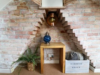 A wooden table with a blue lamp in a brick alcove with a plant in a basket and a crate with cushions at Harbour Reach Brewers Quay Harbour