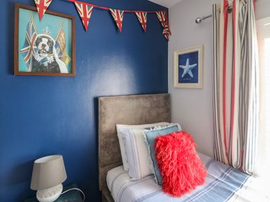 A bedroom with a bed with pillows next to a window with striped curtains and a blue wall with a framed picture of a dog wearing a crown and Union Jack bunting at Harbour Reach Brewers Quay Harbour