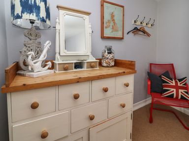 A wooden dresser with a mirror a lamp and a jar of shells a red chair with pillows and coat hooks with hangers on a wall at Harbour Reach in Brewers Quay Harbour