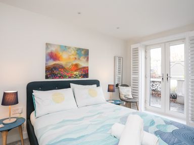A bedroom with a bed a painting bedside tables lamps chair and glass doors at Steps Away in Benllech