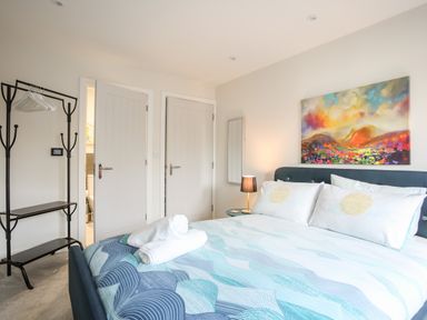 A bedroom with a bed with folded towels on it a colorful painting above the bed a coat rack with hangers two doors and a bedside table with a lamp at Steps Away in Benllech