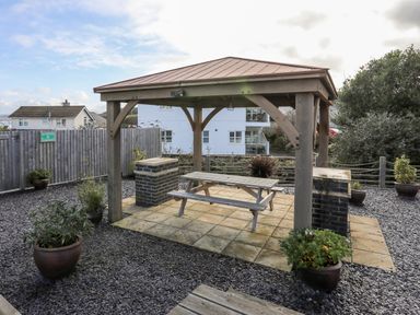 An outdoor wooden gazebo with a picnic table and brick barbeques surrounded by potted plants at Steps Away in Benllech