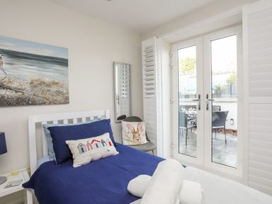 A bedroom with a single bed with blue bedding a chair with a bunny cushion a painting on the wall and glass doors leading outside at Steps Away in Benllech