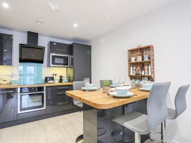 A kitchen with dark cabinets wooden countertop dining table set with bowls plates and glasses and a wall shelf with mugs and containers at Steps Away in Benllech