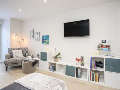 A living room with a patterned armchair a TV on the wall and white shelving units at Steps Away in Benllech