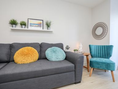 A living room with a gray sofa with yellow and blue round pillows a small wooden side table with decorations and a teal chair at Steps Away Benllech
