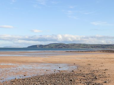A sandy beach with scattered rocks and a view of the sea with distant hills under a blue sky at Steps Away in Benllech