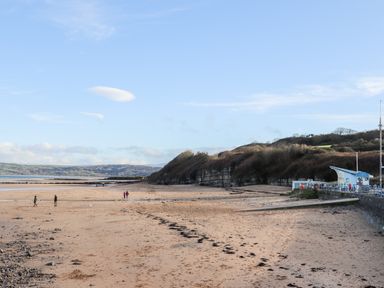 A sandy beach with people walking near a rocky hillside and a small building at Steps Away in Benllech