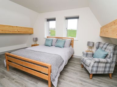 A bedroom with a wooden bed and gray bedding two windows a plaid armchair and two side tables with lamps at Kerry Hill - The Sheepfold in Llanyre near Llandrindod Wells