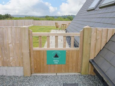 A wooden gate with a green sign saying Kerry Hill leading to a garden with wooden chairs and a table at Kerry Hill - The Sheepfold in Llanyre near Llandrindod Wells