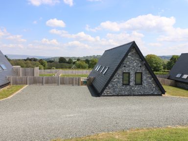 A triangular stone house with black roof tiles surrounded by gravel and wooden fencing with countryside in the background at Kerry Hill - The Sheepfold in Llanyre near Llandrindod Wells