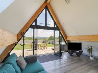 A living room with a teal couch a TV on a stand a potted plant and open glass doors leading to a patio and garden at Kerry Hill - The Sheepfold in Llanyre near Llandrindod Wells