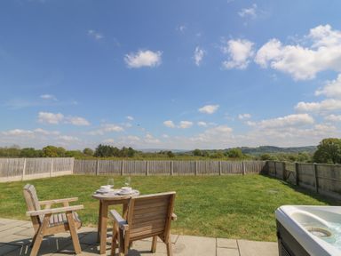 A backyard with a wooden table and chairs set on a patio and a hot tub next to a fenced grass area at Kerry Hill - The Sheepfold in Llanyre near Llandrindod Wells