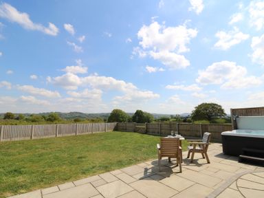 A fenced backyard with grass a hot tub and wooden chairs on a stone patio at Kerry Hill - The Sheepfold in Llanyre near Llandrindod Wells