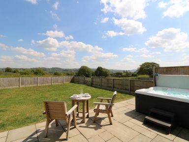 A backyard with wooden chairs and table set on a patio next to a hot tub with a fenced lawn and trees in the background at Kerry Hill - The Sheepfold in Llanyre near Llandrindod Wells