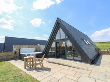 An outdoor patio area with wooden chairs and a table next to a hot tub and an a frame building with large glass doors at Kerry Hill - The Sheepfold in Llanyre near Llandrindod Wells