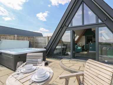 An outdoor patio with a hot tub wooden table and chairs set with dishes and glassware next to a glass door leading to a living area at Kerry Hill - The Sheepfold in Llanyre near Llandrindod Wells