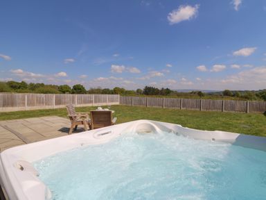 An outdoor jacuzzi with bubbling water and wooden chairs and table on a patio in a fenced grassy yard at Kerry Hill - The Sheepfold Llanyre near Llandrindod Wells