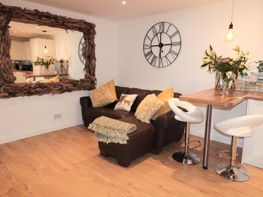 A living area with a brown leather sofa and cushions next to a wooden breakfast bar with two white stools and a large wall clock at Swale Cottage in Beadnell