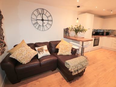 A living room with a brown leather sofa and cushions next to a kitchen with white cabinets at Swale Cottage in Beadnell