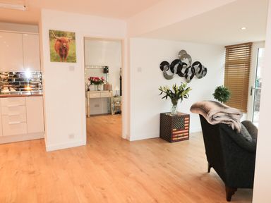 A room with wooden flooring a black armchair with a blanket an american flag patterned side table with flowers a wall art a door with wooden blinds and a kitchen area with white cabinets at Swale Cottage in Beadnell