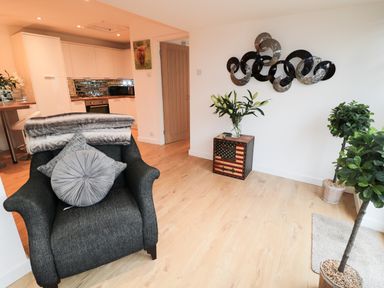 A living area with a dark armchair, pillows, a blanket, a decorative metal wall piece, a small table with flowers, two potted plants and a kitchen in the background at Swale Cottage in Beadnell