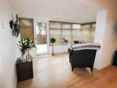 A sitting area with a dark armchair a side table with flowers two potted plants and a glass door with blinds at Swale Cottage in Beadnell
