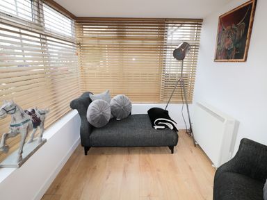 A corner room with a chaise lounge, wooden blinds on windows, a floor lamp, a white radiator, a horse sculpture, and a colorful painting on the wall at Swale Cottage in Beadnell