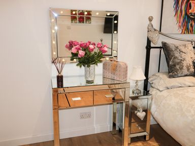 A mirrored dressing table with pink roses in a vase a reed diffuser and a floral bag next to a mirrored bedside table with a lamp and a bed at Swale Cottage in Beadnell