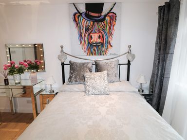 A bedroom with a bed with decorative pillows bedside tables with lamps a mirrored vanity table with flowers dark curtains and a colorful cow painting above the bed at Swale Cottage in Beadnell