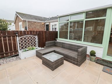 An outdoor patio with a wicker corner sofa and table next to a green framed window at Swale Cottage in Beadnell