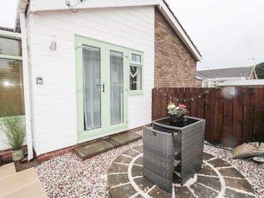 A small outdoor patio area with a wicker table and chair set on circular stone paving outside a white building with pale green doors and window at Swale Cottage in Beadnell