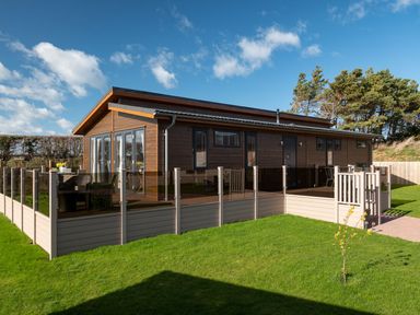 A wooden cabin with glass railing on a deck surrounded by green grass and trees at Lobster Lodge in Runswick Bay