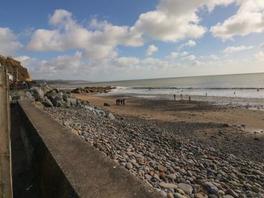 A rocky beach with people walking near the shore and a concrete wall in the foreground at Cwtch Lodge 42 in Stepaside near Wisemans Bridge