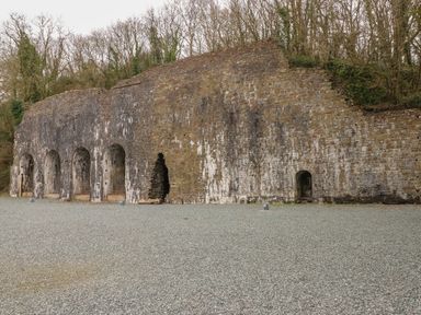 A large stone wall with multiple arched openings on a gravel ground in front of trees at Cwtch Lodge 42 in Stepaside near Wisemans Bridge