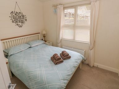 A bedroom with a bed with blue patterned bedding and towels on top a window with blinds and curtains and a lamp on a bedside table at Cwtch Lodge 42 Stepaside near Wisemans Bridge