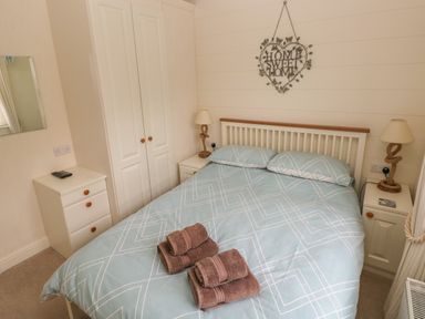A bedroom with a double bed with blue patterned bedding and folded towels on top bedside tables with lamps and a wardrobe at Cwtch Lodge 42 in Stepaside near Wisemans Bridge