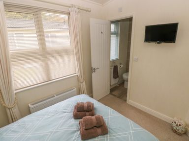 A bedroom with a bed towel sets on it a window with blinds an open door leading to a bathroom and a wall-mounted television at Cwtch Lodge 42 in Stepaside near Wisemans Bridge