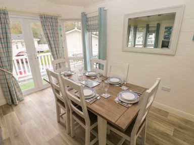 A dining area with a wooden table set for six by windows with curtains at Cwtch Lodge 42 Stepaside near Wisemans Bridge