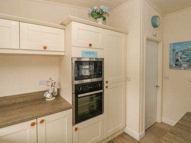 A kitchen with a built-in microwave and oven a countertop with mugs on a stand a clock on the wall and a door at Cwtch Lodge 42 Stepaside near Wisemans Bridge