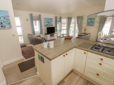 A kitchen area with a stove and counter overlooking a living room with couches a TV and windows at Cwtch Lodge 42 Stepaside near Wisemans Bridge