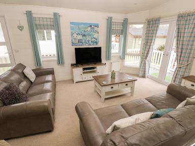 A living room with two brown sofas a coffee table a TV on a stand and windows with curtains at Cwtch Lodge 42 Stepaside near Wisemans Bridge