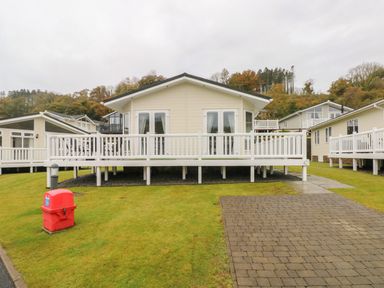 A beige lodge with white railings and multiple windows on a grassy plot next to a paved driveway at Cwtch Lodge 42 Stepaside near Wisemans Bridge