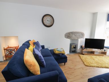 A living room with a blue sofa and yellow cushions a clock on the wall a television on a wooden stand and a yellow rug at Bryn Derfel in Borth-y-Gest near Porthmadog