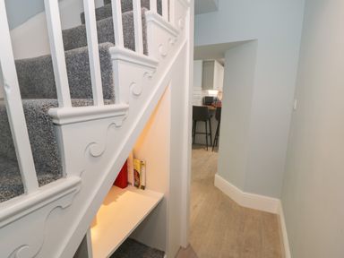 A hallway with carpeted stairs and a small bookshelf under the stairs next to a kitchen with bar stools at Bryn Derfel in Borth-y-Gest near Porthmadog