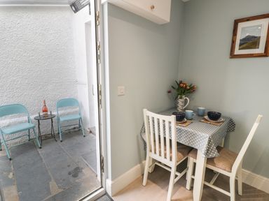 A small dining area with a table covered in a polka dot tablecloth set with bowls and mugs next to an open door leading to an outdoor space with two blue chairs and a small table holding a bottle and glasses at Bryn Derfel in Borth-y-Gest near Porthmadog