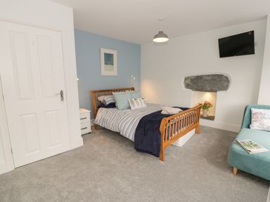 A bedroom with a wooden bed with striped bedding two bedside tables a blue accent wall a wall-mounted TV a small alcove with a stone feature and a vase of flowers at Bryn Derfel Borth-y-Gest near Porthmadog